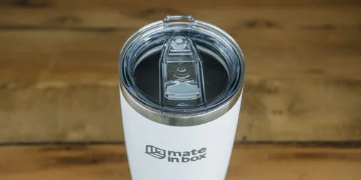 Copo Térmico em primeiro plano em cima de uma mesa de madeira. Foto mostra o copo em corpo inteiro, com tampa para bebidas quentes ou geladas. A tampa é abre fecha, facilitando assim viagens com borrachas de vedação. Tampa é em acrílico.