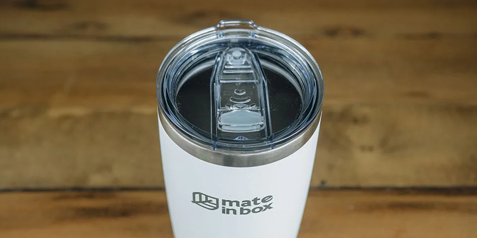 Copo Térmico em primeiro plano em cima de uma mesa de madeira. Foto mostra o copo em corpo inteiro, com tampa para bebidas quentes ou geladas. A tampa é abre fecha, facilitando assim viagens com borrachas de vedação. Tampa é em acrílico.