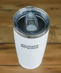 Copo Térmico em primeiro plano em cima de uma mesa de madeira. Foto mostra o copo em corpo inteiro, com tampa para bebidas quentes ou geladas. A tampa é abre fecha, facilitando assim viagens com borrachas de vedação. Tampa é em acrílico.