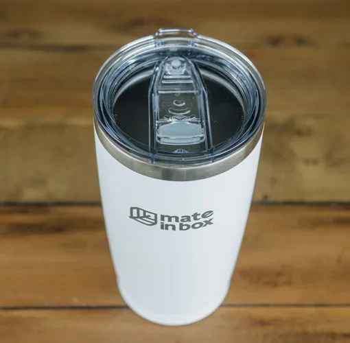 Copo Térmico em primeiro plano em cima de uma mesa de madeira. Foto mostra o copo em corpo inteiro, com tampa para bebidas quentes ou geladas. A tampa é abre fecha, facilitando assim viagens com borrachas de vedação. Tampa é em acrílico.