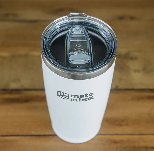 Copo Térmico em primeiro plano em cima de uma mesa de madeira. Foto mostra o copo em corpo inteiro, com tampa para bebidas quentes ou geladas. A tampa é abre fecha, facilitando assim viagens com borrachas de vedação. Tampa é em acrílico.